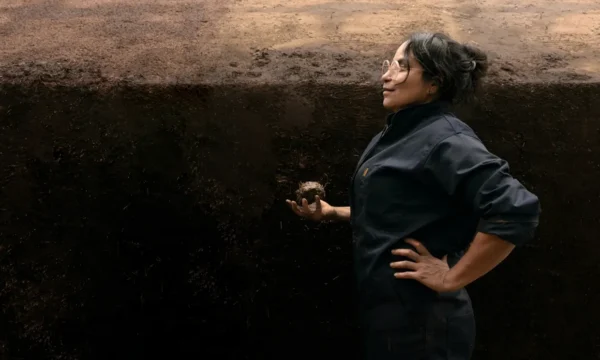 Artist Delcy Morelos holding a lump of earth standing next to a huge earthen sculpture