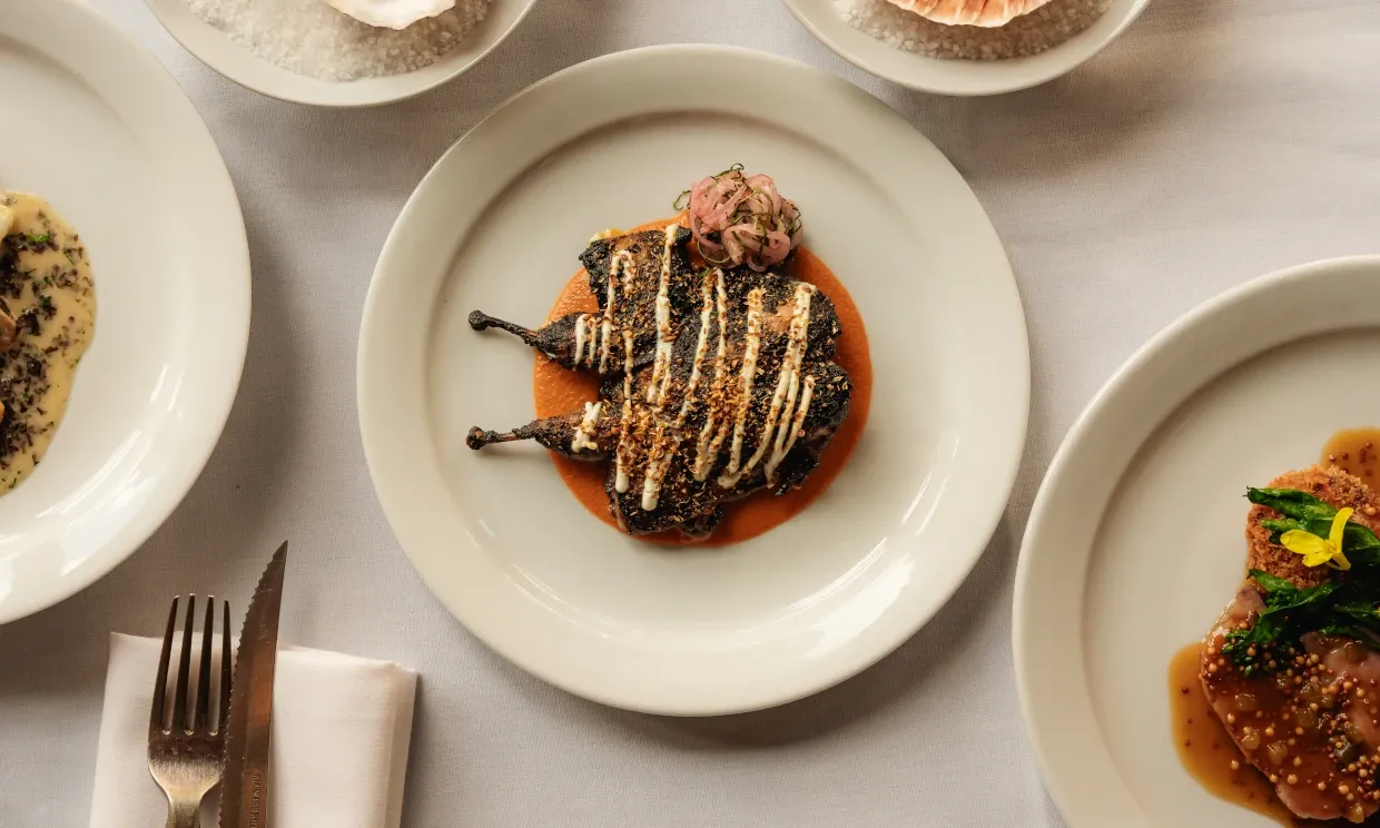 A small quail sits on a place, with orange sauce and some pickles.