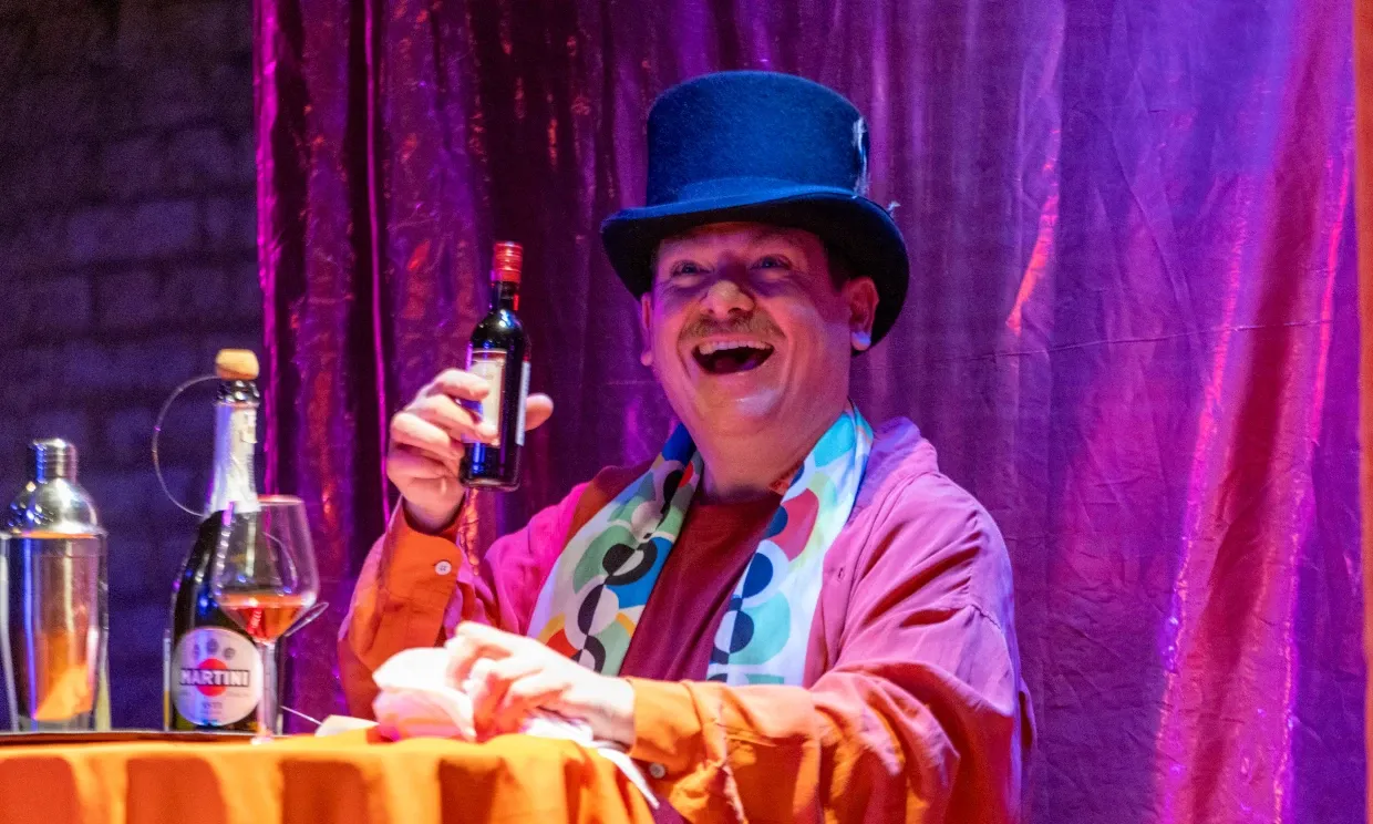 A production still from A Fine Idea. Kevin is sat at a table in a top hat, drinking Martini with a huge smile on his face