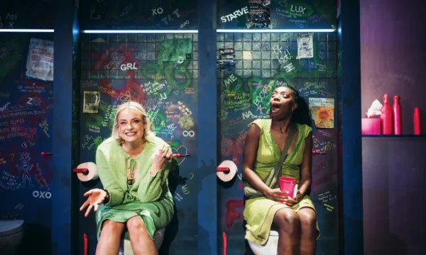 Two women sit in toilet cubicles, lit by stage lights