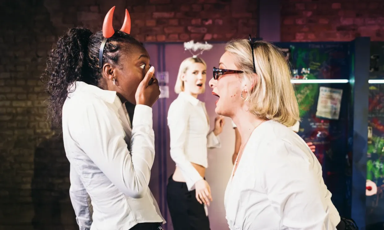 Three women talking in a bathroom, one is wearing devil horns and the other an angel halo.