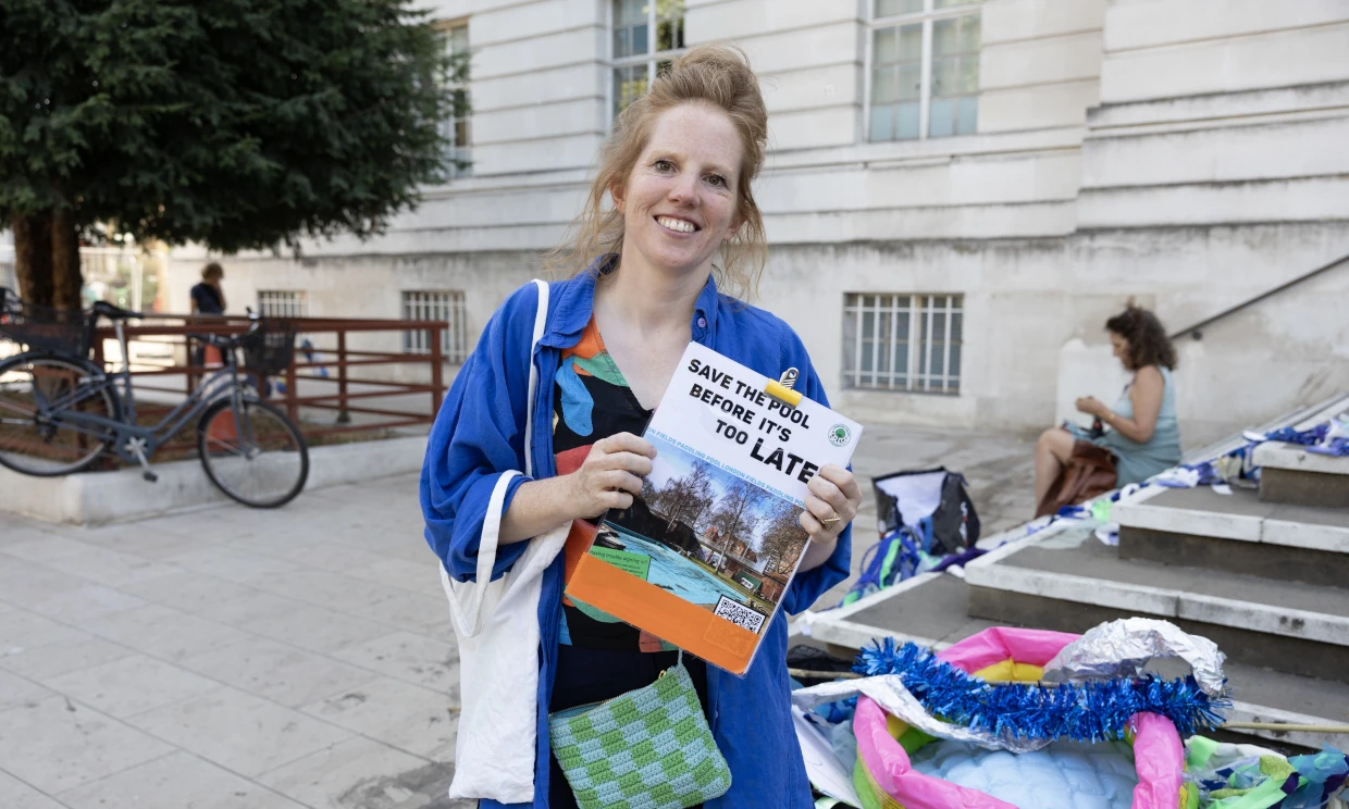 A woman stands at the protest with a clipboard sign that reads "Save the pool before it's too late".