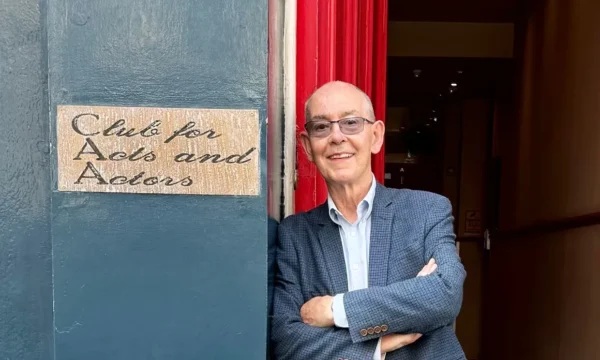 Eden stands outside a theatre, leaning against a wall with a sign on that reads "Club for Acts and Actors"