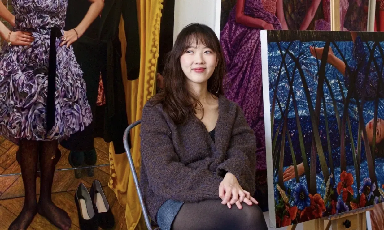 Photo of a young woman sat beside some of her paintings, smiling.