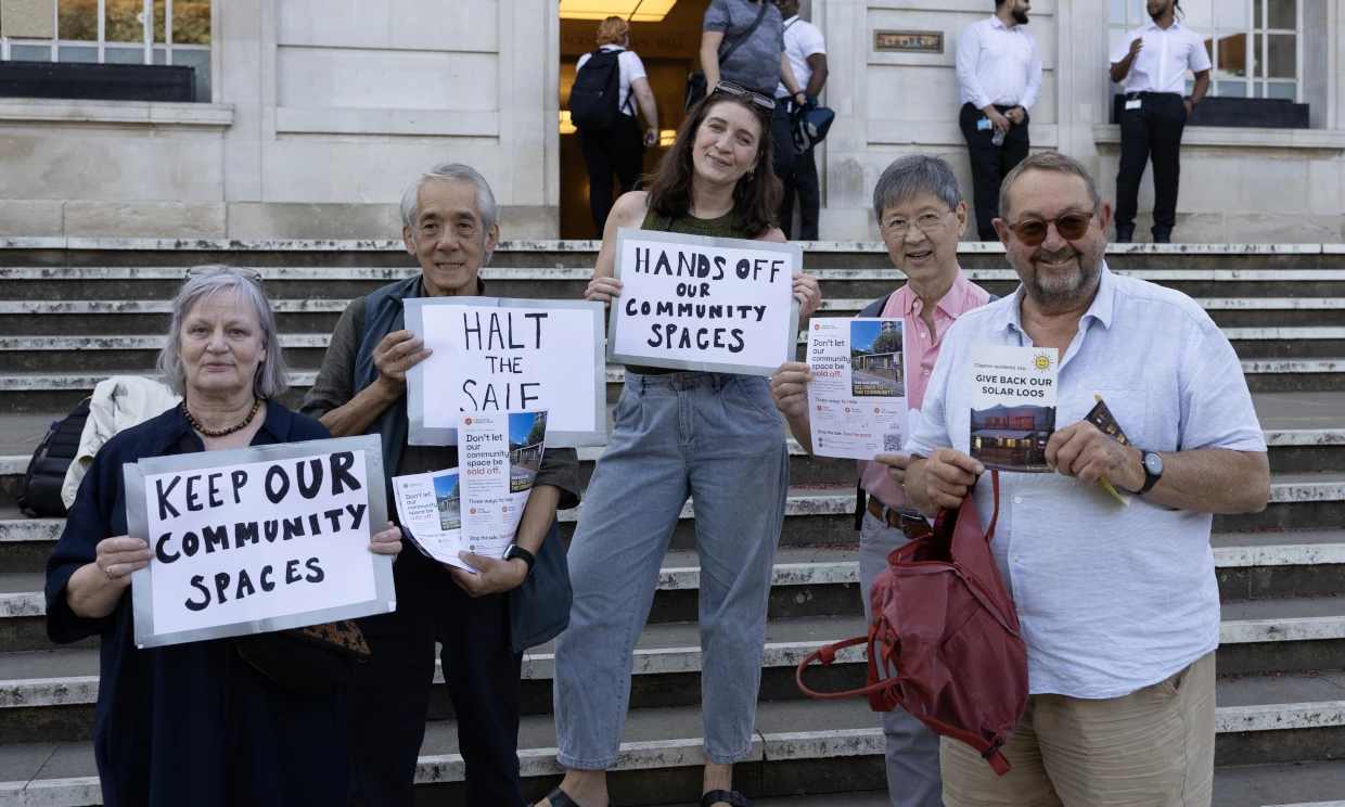 A small group of protestors gather with signs. One reads "Hands off our community spaces".