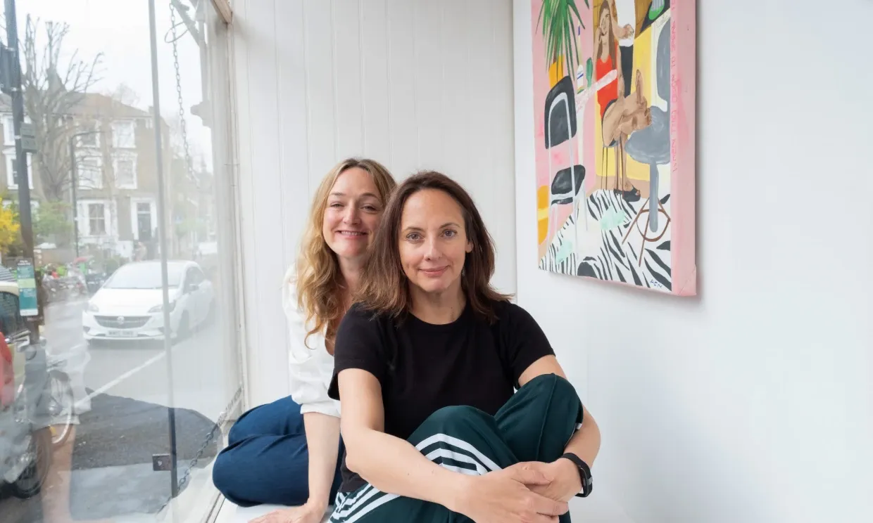 Two women sat next to a canvas painting smiling.