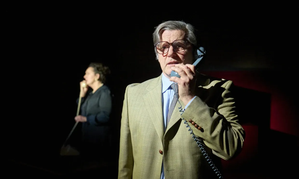 A man in a suit talking on a cord phone, behind him is a woman on the receiving end of the phone. The stage is dark, with only the two figures lit