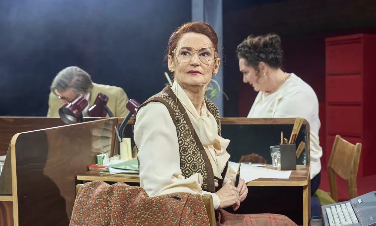 A woman in 1950s attire sits at a desk, beside her there are two other people working