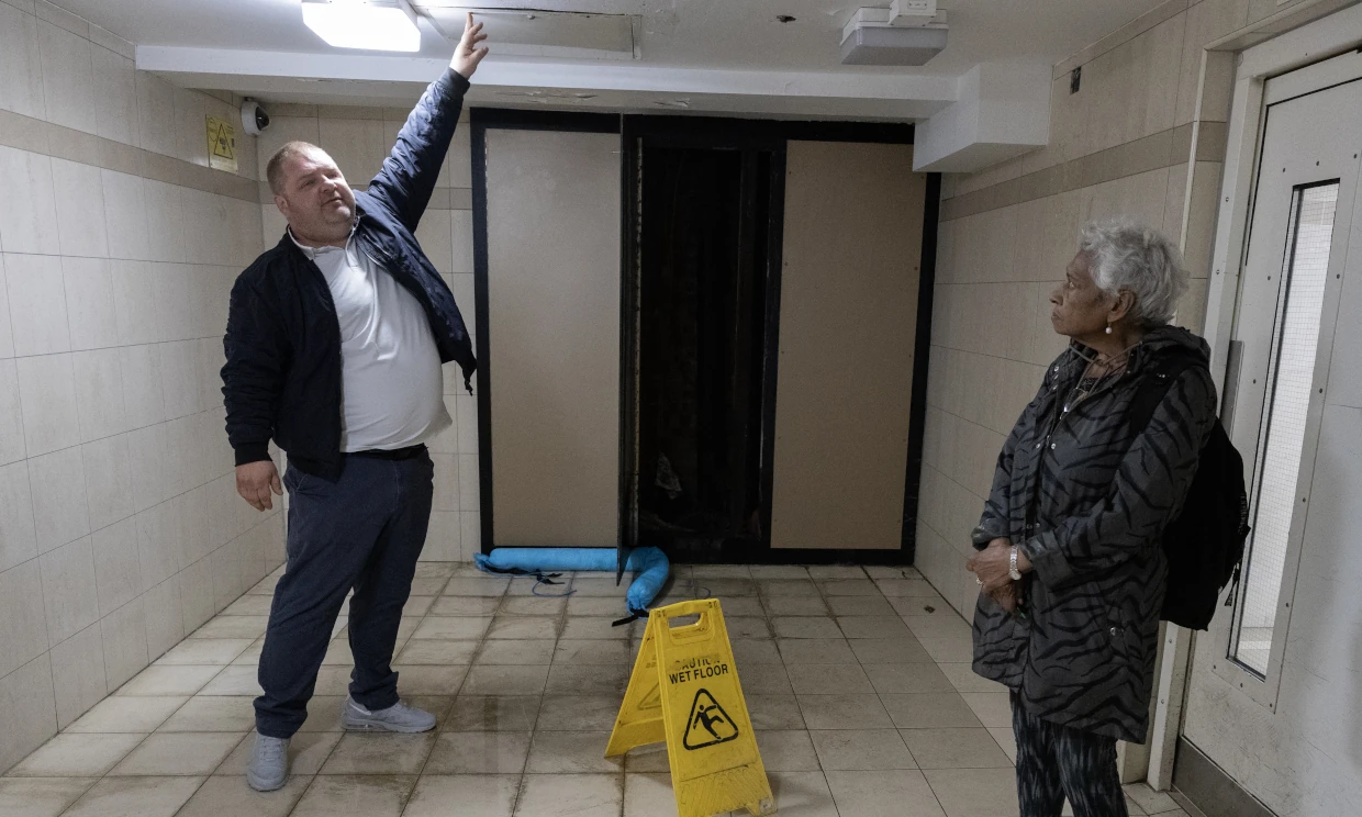 A man is standing in a puddle of water inside, he is touching a leak on the ceiling above.