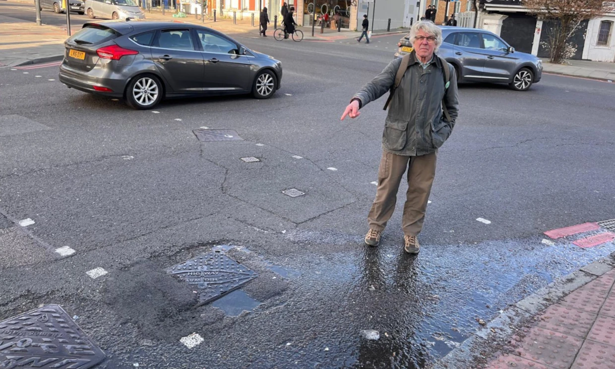 Reiner Tegtmeyer points at a leaking pothole in the road, water leaks down the curb.