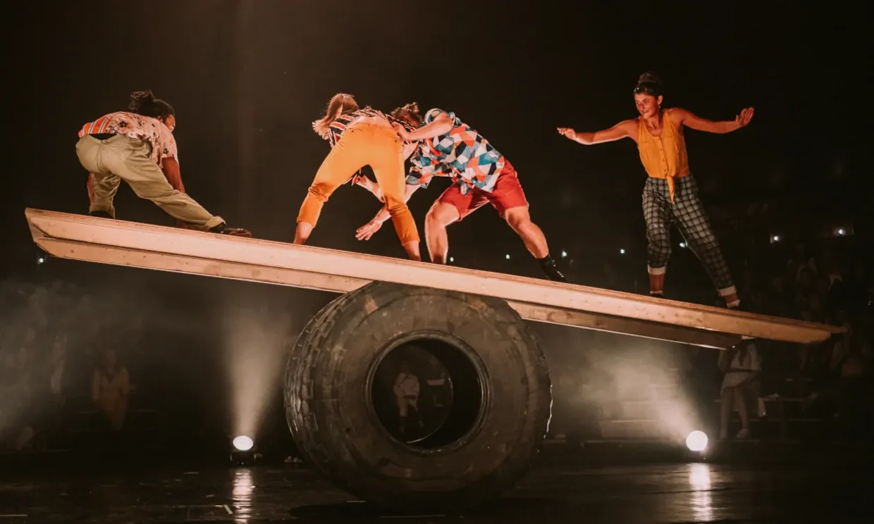 4 people balance on a wooden plank rolling on a huge tyre.
