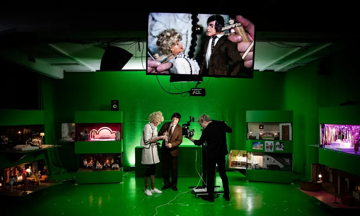 A zoomed out shot of the studio, which features several tiny sets. Three people stand in the middle of the studio, two working the puppets and one cameraman.