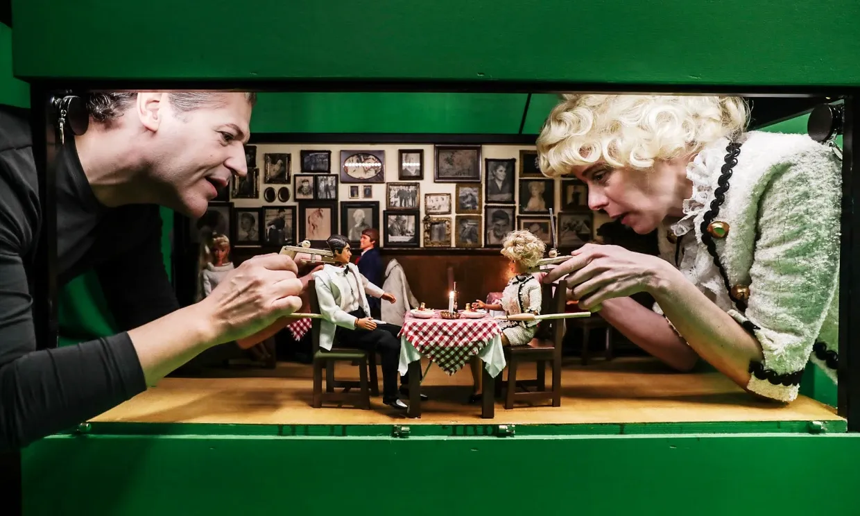 A more zoomed out shot, showing two puppeteers working two puppets sat at a dining table