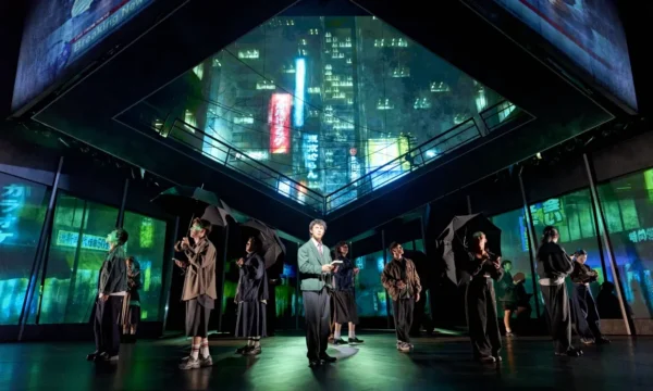 An ensemble on stage. A young man is lit up in the centre, people stand around him in black clothes with umbrella's under a futuristic skyline.