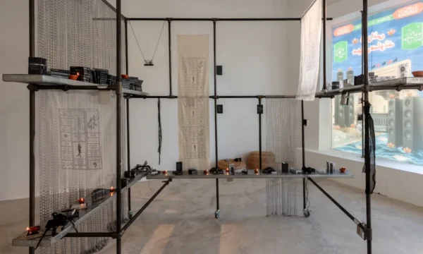 Art installation that is a series of metal poles forming a structure with shelves. On the shelves are cassettes and other items arranged with candles in-between.